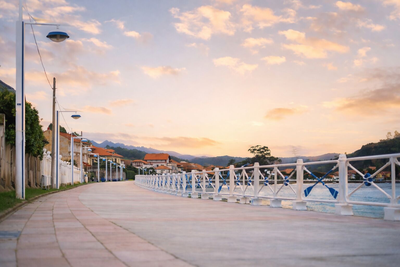 A waterfront promenade with a white railing, street lamps, and a row of buildings with red roofs.