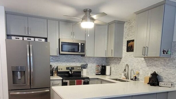 A modern kitchen with grey cabinets, a stainless steel refrigerator, a microwave, and a built-in oven. There is a small trash bin and a bar stool area with three stools.
