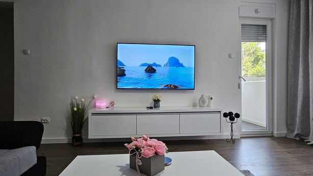 Ein modernes Wohnzimmer mit einem an der Wand montierten Flachbildfernseher, einem weißen Couchtisch mit floraler Dekoration, einem dunkelgrauen Sofa und einem schlichten, weißen Schrank.
