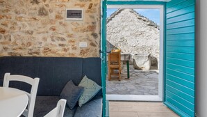 Une pièce avec un mur en pierre, une porte de couleur turquoise, un canapé foncé, une table blanche et une vue sur une structure en pierre à l’extérieur.
