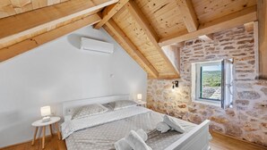 Une chambre avec un plafond en pente, des murs en pierre, un lit avec une literie blanche, une petite table de chevet et une fenêtre donnant sur un beau paysage.