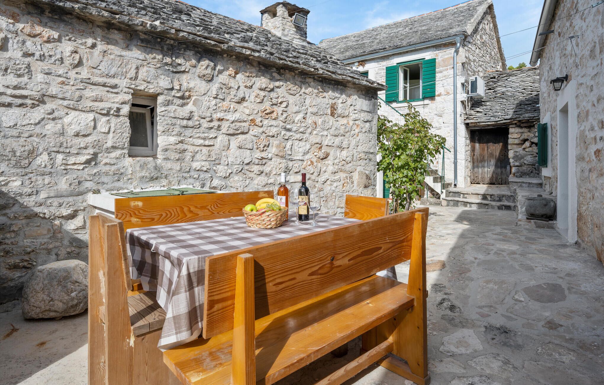 Un bâtiment en pierre avec une table et des chaises en bois, un panier de fruits et une bouteille de vin.