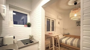 A compact kitchen with a sink, a window, and a small table with a chair. There is a bed with a striped blanket and a hanging light fixture.
