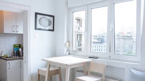 A small, bright kitchen with white cabinets, a white table, and two chairs. There is a window with a view of buildings outside.
