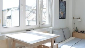 A bright room with a large window, a white table, a bed with pillows, a wall-mounted lamp, and a painting.