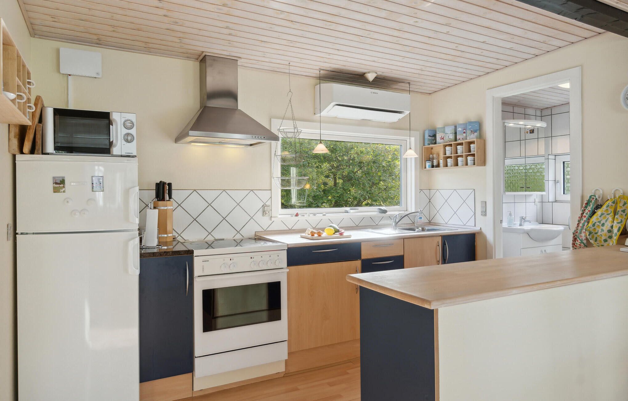 Une cuisine moderne équipée d’un four encastré, d’un four à micro-ondes et d’une cuisinière. Une fenêtre laisse entrer la lumière naturelle et offre une vue sur la verdure environnante.