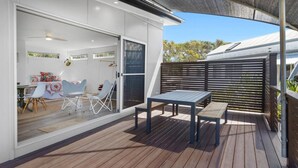 Un patio moderno al aire libre con terraza de madera, un área de estar cubierta y un ventanal corredizo que da a un espacio interior con una mesa y sillas de comedor.