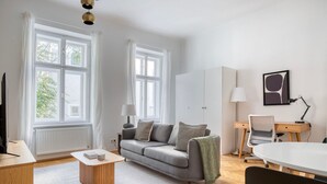En moderne stue med en grå sofa, et træbord, et hvidt spisebord og et skrivebord med en stol. To vinduer med hvide gardiner, en radiator og en lampe på skrivebordet.