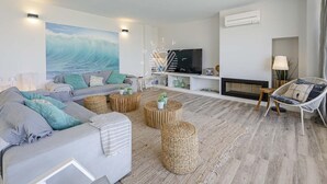 A modern living room with a grey sofa, round wooden side tables, a flat-screen TV, and a large wave painting on the wall. - Casa del Mar · Pool & Sea Views · 10 Guests (Santa Margalida - Can Picafort/Son Serra Marina)