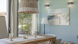 A dining area with a wooden table, white chairs, a wicker pendant light, and a beach-themed painting on the wall. - Casa del Mar · Pool & Sea Views · 10 Guests (Santa Margalida - Can Picafort/Son Serra Marina)