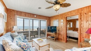 En træhytte med stor skydedør til stranden, ventilator i loftet, fjernsyn og en sofa med mønstrede puder.