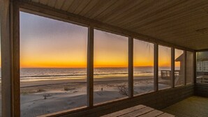 En træterrasse med en bænk, store vinduer med udsigt over stranden og havet i solnedgangen.