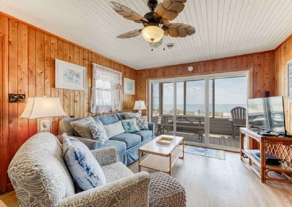 Un salon chaleureux avec un canapé bleu, des fauteuils à motifs, un ventilateur de plafond en bois et une vue sur l’extérieur, visible à travers une porte en verre.