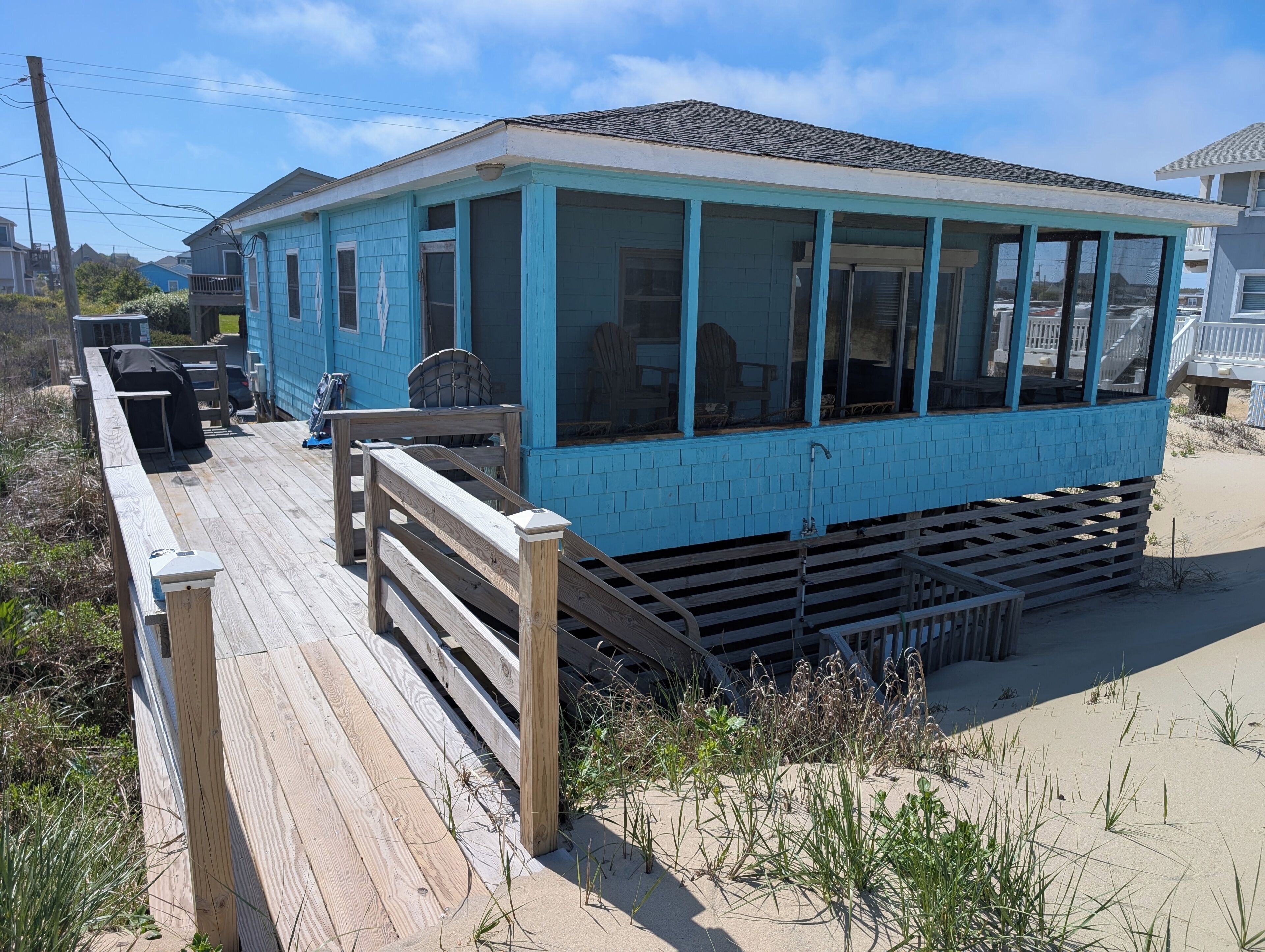 A blue building with a wooden deck, a grill, and a sandy beach in the background.