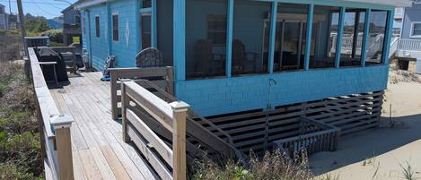 A blue building with a wooden deck, a grill, and a sandy beach in the background.