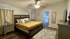 Quarto com cama, mesas de cabeceira, escrivaninha, ventilador de teto e vista para o banheiro.