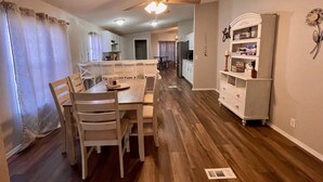 Uma área de refeições com mesa e cadeiras de madeira, um ventilador de teto e uma cozinha ao fundo.