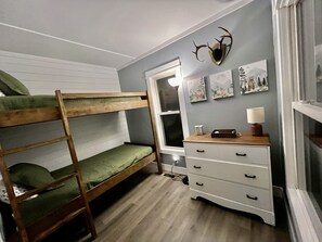 Un lit superposé avec une literie verte, une commode à tiroirs, une lampe et des décorations murales.