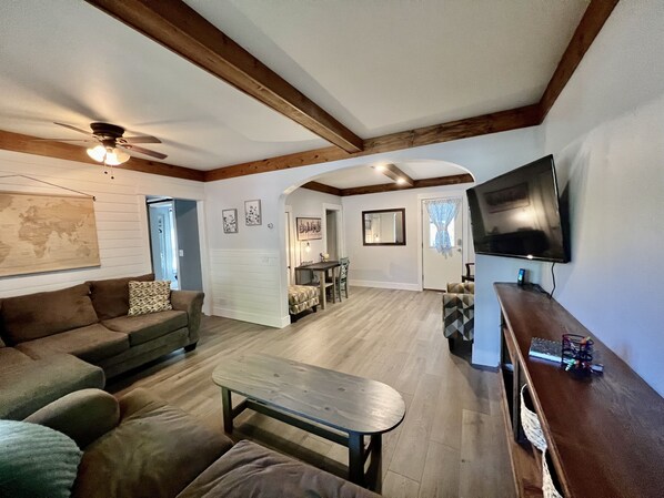 Un vaste salon doté d’un plafond en bois, d’un grand téléviseur à écran plat fixé au mur, d’un canapé, d’une table basse et d’un coin repas avec une table et des chaises.