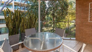 A round glass table with wicker chairs, a view of a building, and tall plants. - Oceancrest 11 on Forster main beach. (Forster)