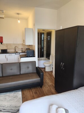 A compact hotel room with a sofa, a wardrobe, a small kitchenette, a toilet, and a shower area. - 1 Bed Flat - Sleeps 3 - Parking - Wifi (Greenhithe)