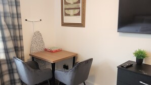 Ein kleiner, ordentlich eingerichteter Raum mit einem Holztisch, zwei grauen Stühlen, einem schwarzen Schrank, einem gerahmten Kunstwerk und einem Fernseher an der Wand.