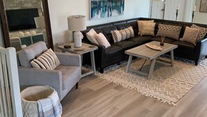 Un salon moderne avec un canapé gris, une table basse et un téléviseur. On y trouve des coussins décoratifs et un tapis sur le sol.