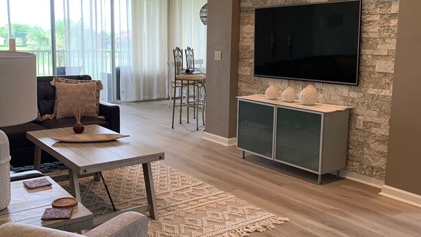 Un salon moderne avec un mur d’accent en pierre, un téléviseur à écran plat, un coin repas avec des chaises et une grande fenêtre avec des rideaux.