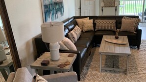 Un salon avec un canapé d’angle, une table basse et une lampe. Un tableau est accroché au mur, et une porte donne sur un balcon.