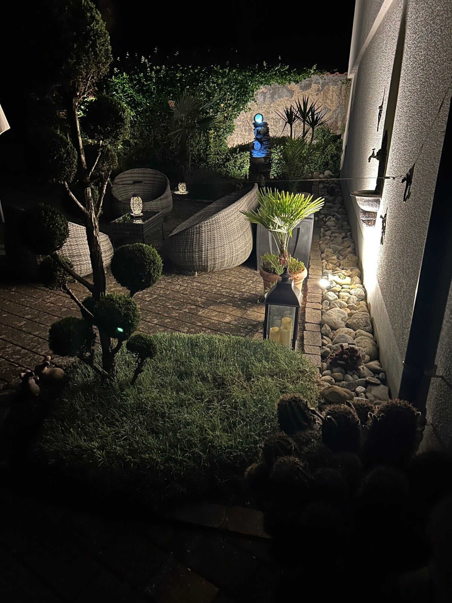 A nighttime outdoor area with a stone pathway, a large woven chair, a potted plant, and a wall-mounted light fixture.