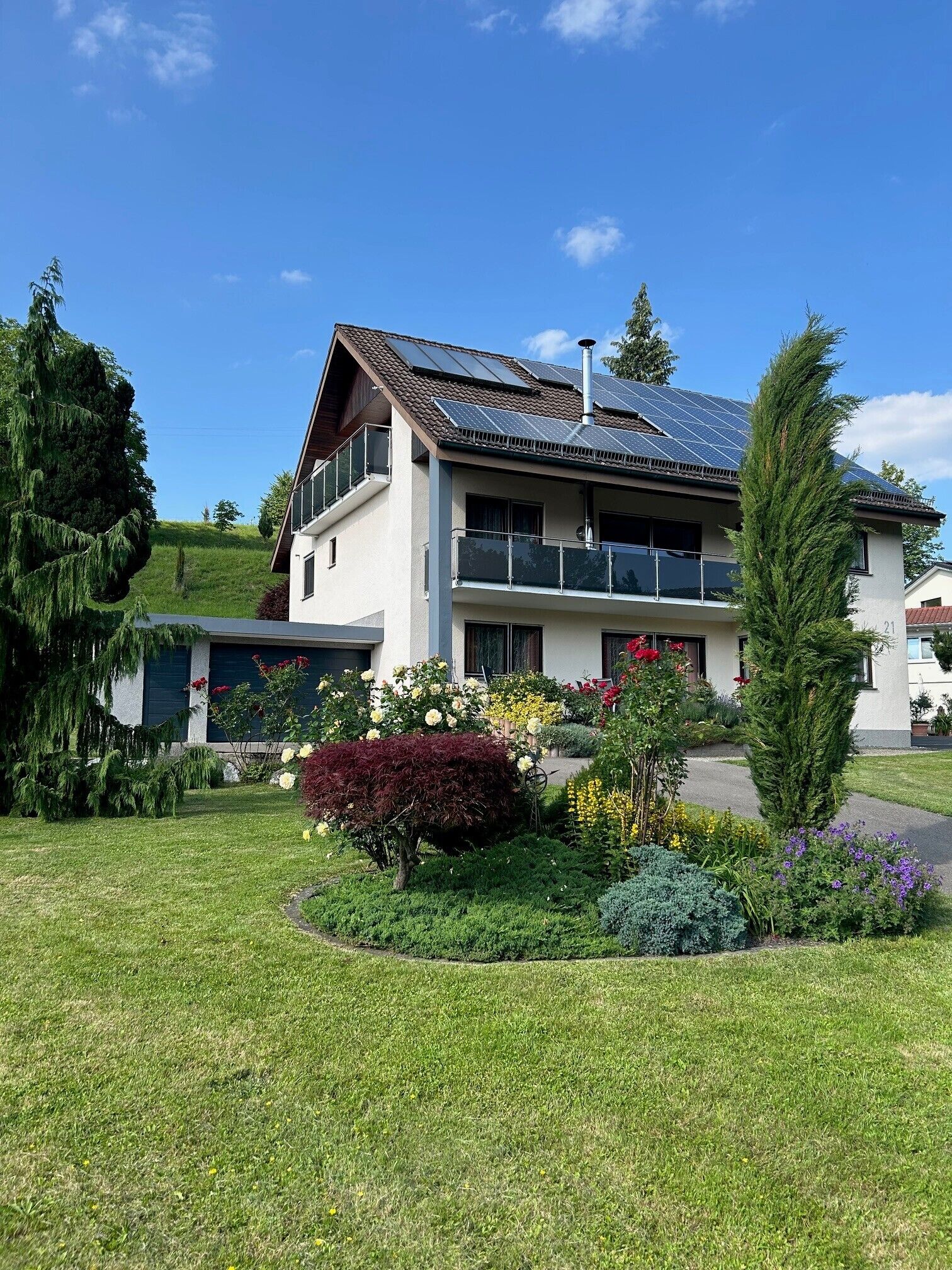 A two-story house with a balcony, solar panels on the roof, and a well-maintained garden with various plants and flowers.