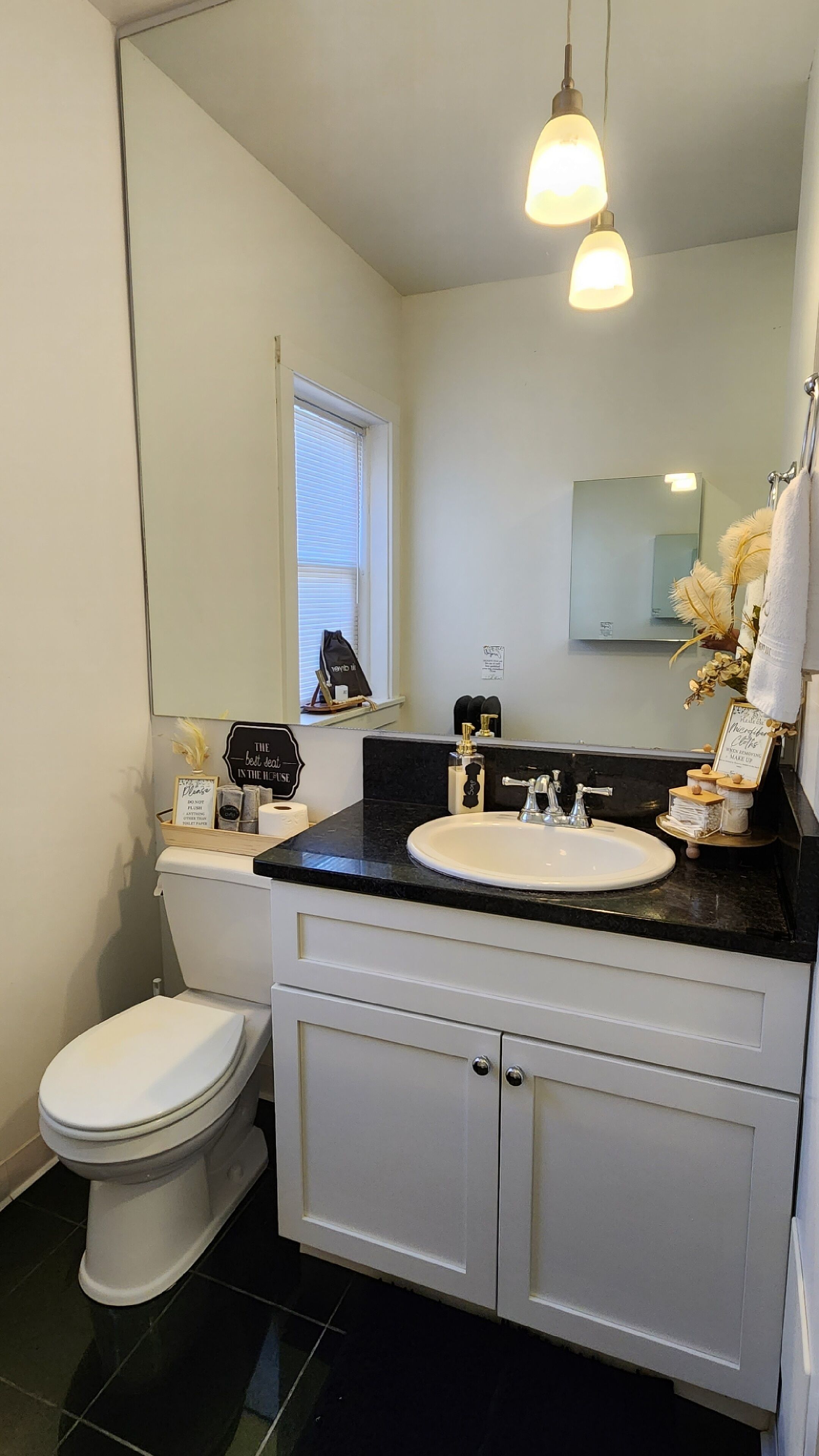 Un baño con inodoro blanco, lavabo negro y espejo sobre el lavabo. Hay una ventana con persianas, un perchero y una señal en la pared.