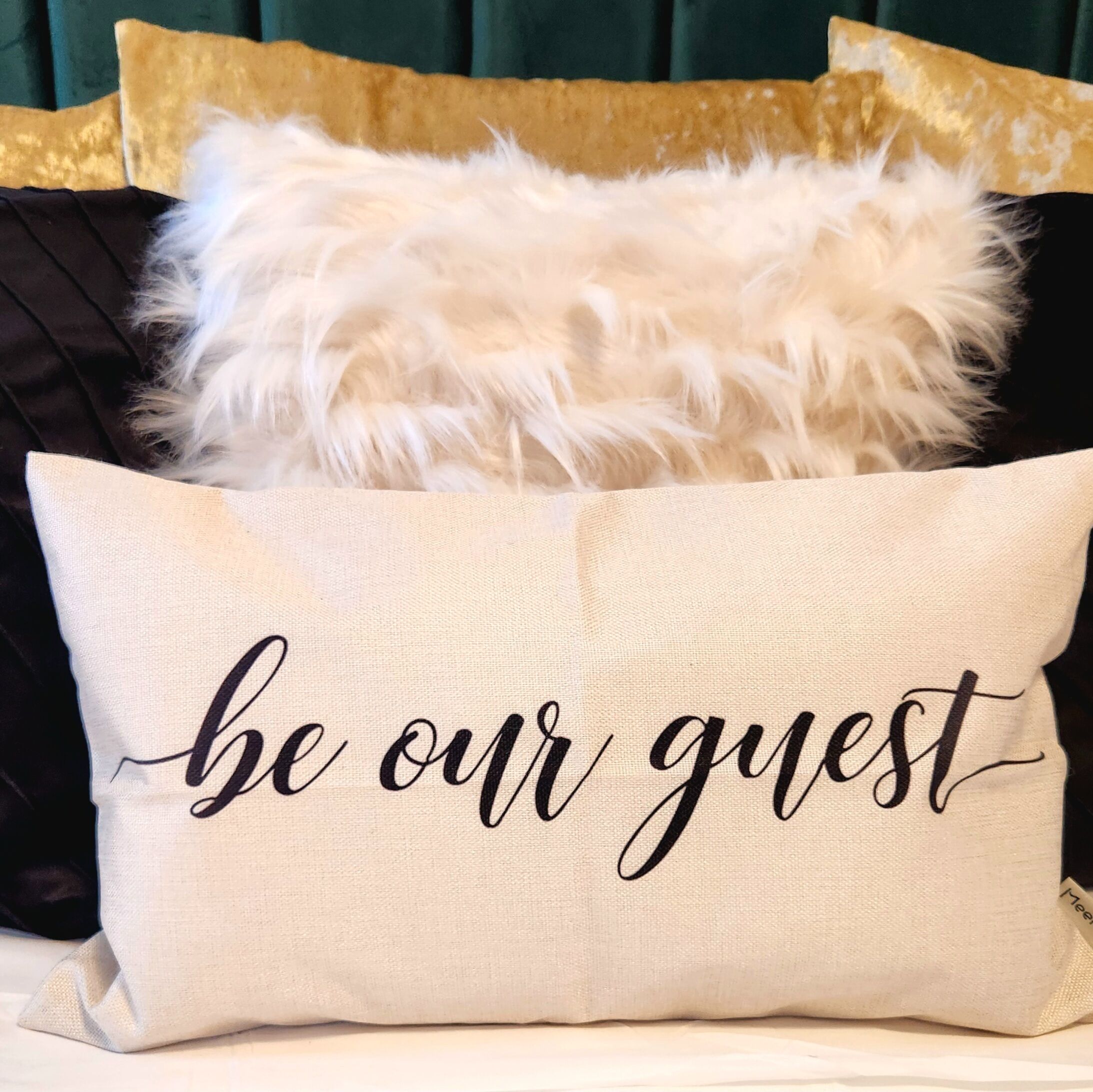 A beige pillow with the text "be our guest" on a bed with a black and gold pillow.