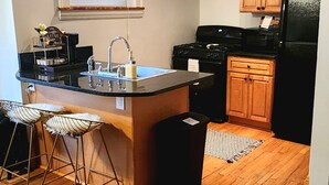 A kitchen with wooden cabinets, a black refrigerator, a stainless steel sink, and a bar stool. - Chic Coventry — Enjoy 10% off 2+ Nights +Late Check Out  (Cleveland Heights)