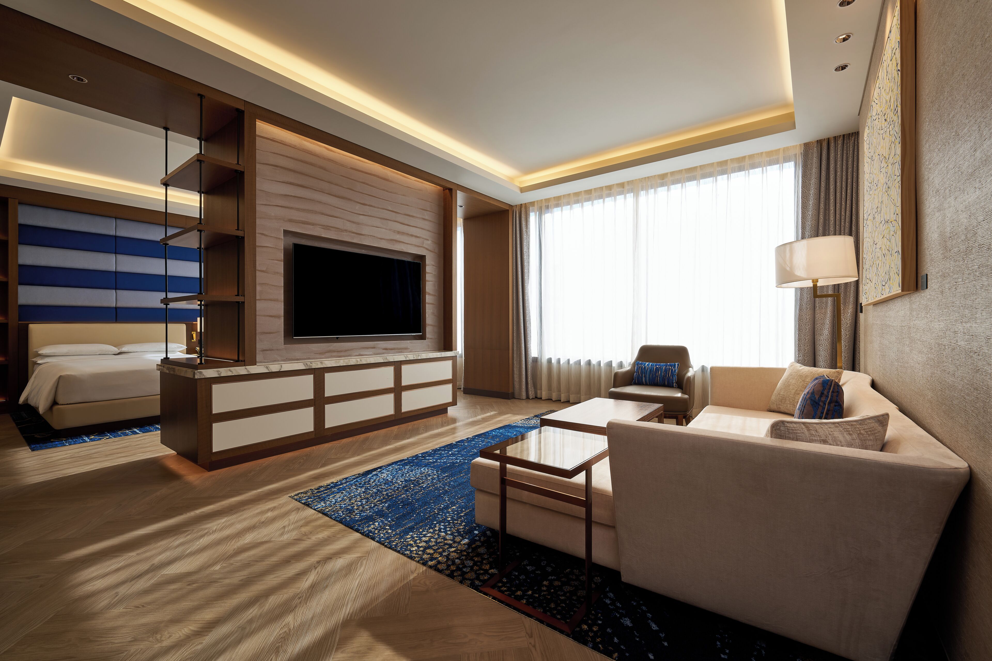 A modern hotel room with a flat-screen TV, a sofa, a coffee table, a bed, and a large window with curtains.