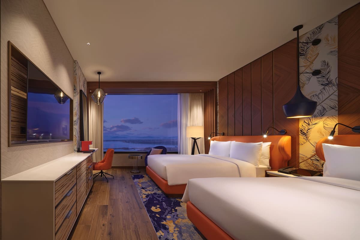 A hotel room with two beds, a desk, a chair, a view of the water, and a large window.