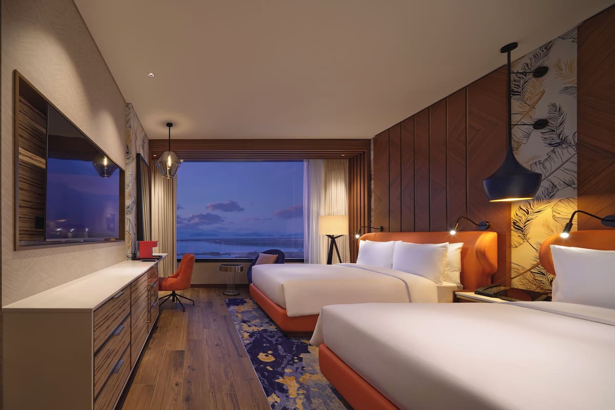 A hotel room with two beds, a desk, a chair, a view of the water, and a patterned wall.