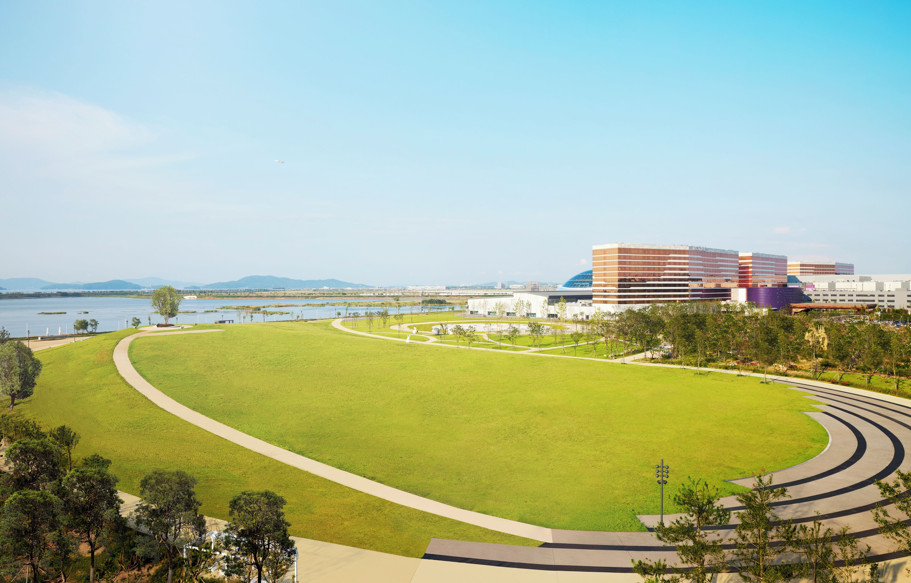A large, open grassy area with a curved pathway leading to a modern building complex by a body of water.