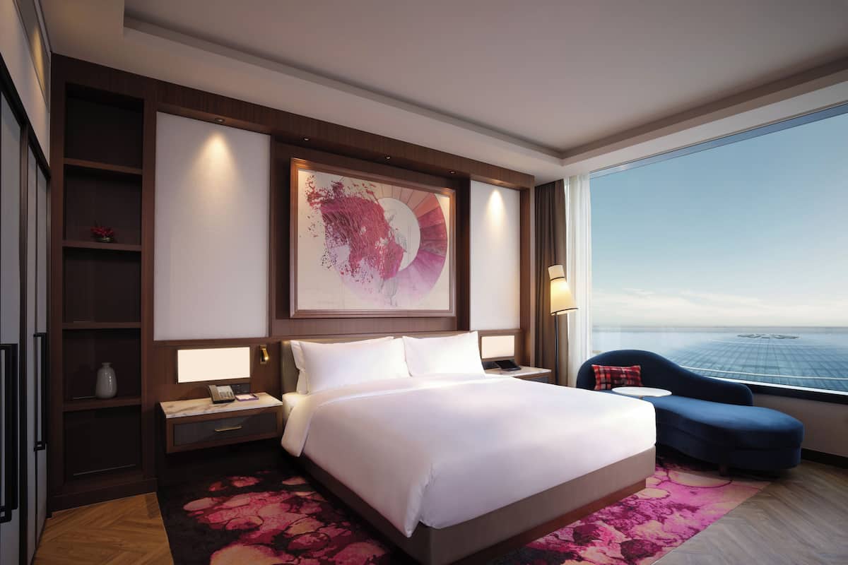 A hotel room with a large bed, a desk with a lamp, a chair, a large window with a view, and a decorative wall piece.