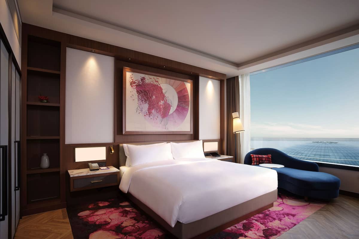 A modern hotel room with a large bed, a desk with a lamp, a chair, and a view of the ocean.