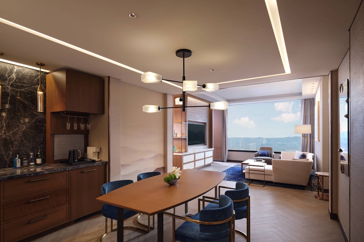 A modern hotel room with a dining area, a seating area with a view, and a kitchen with marble backsplash.