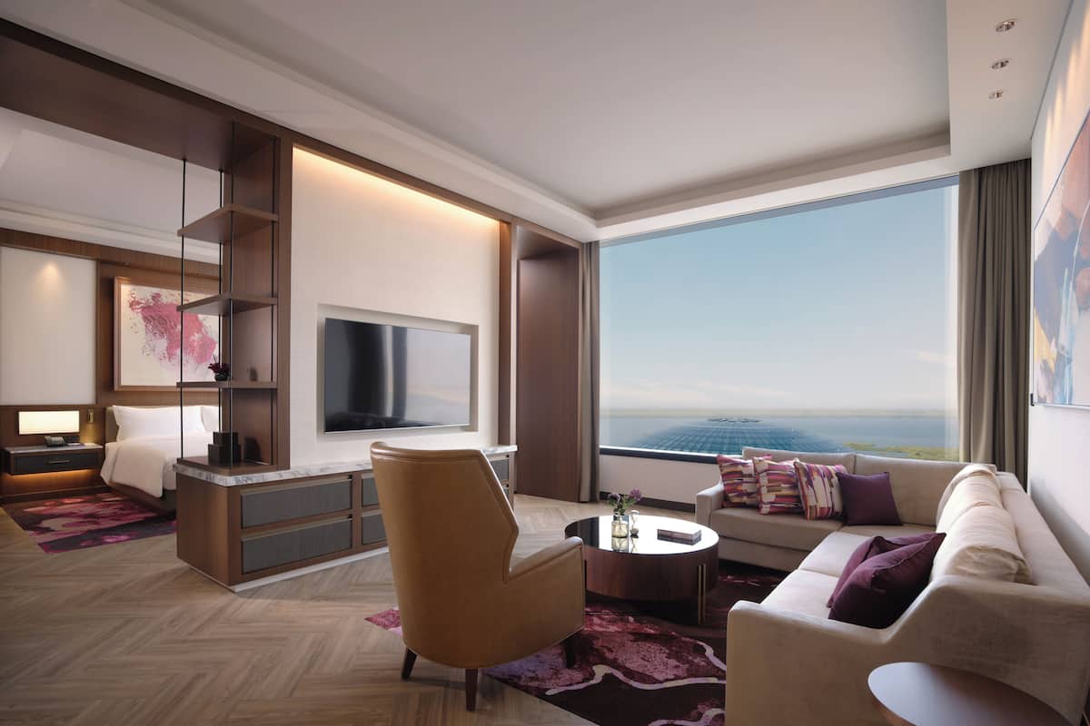 A modern hotel room with a large window, a flat-screen TV, a sofa, a chair, and a small table.