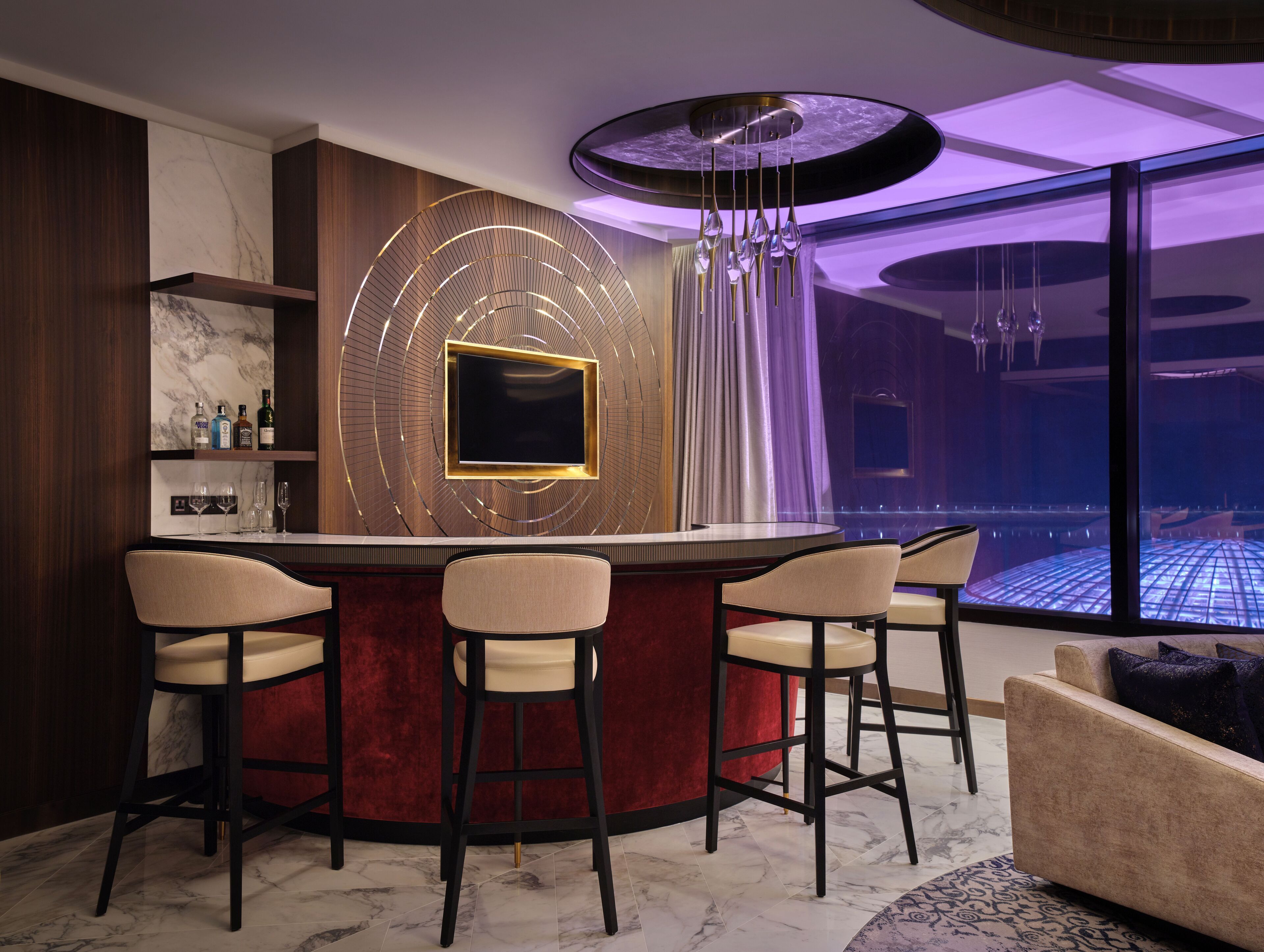 A modern bar area with a circular bar, four bar stools, a TV mounted on a decorative wall, and a view of a pool through large windows.