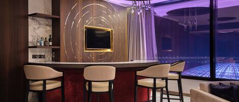 A modern bar area with a circular bar, four bar stools, a TV mounted on a decorative wall, and a view of a pool through large windows.