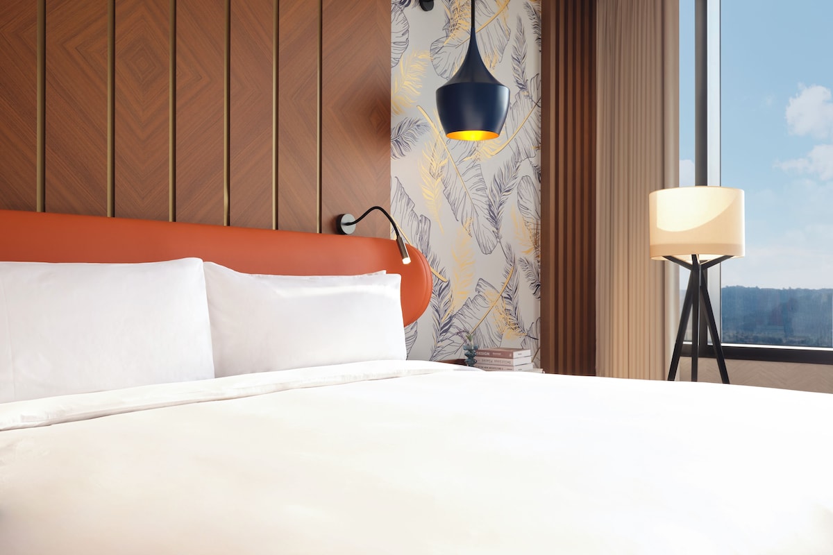 A hotel room with a bed, a wall-mounted lamp, and a view of the outdoors.