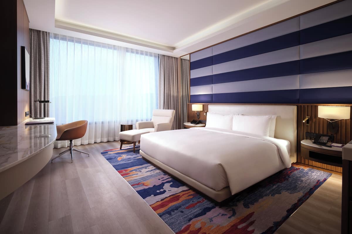 A modern hotel room with a large bed, a desk, a chair, and a view of the city through large windows.