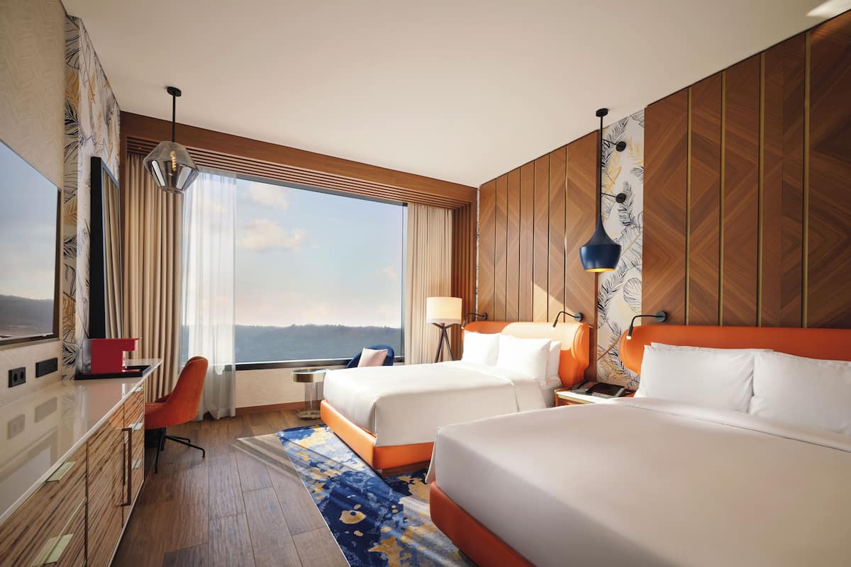 A hotel room with two beds, a desk, a chair, a large window with a view, and a patterned wall.