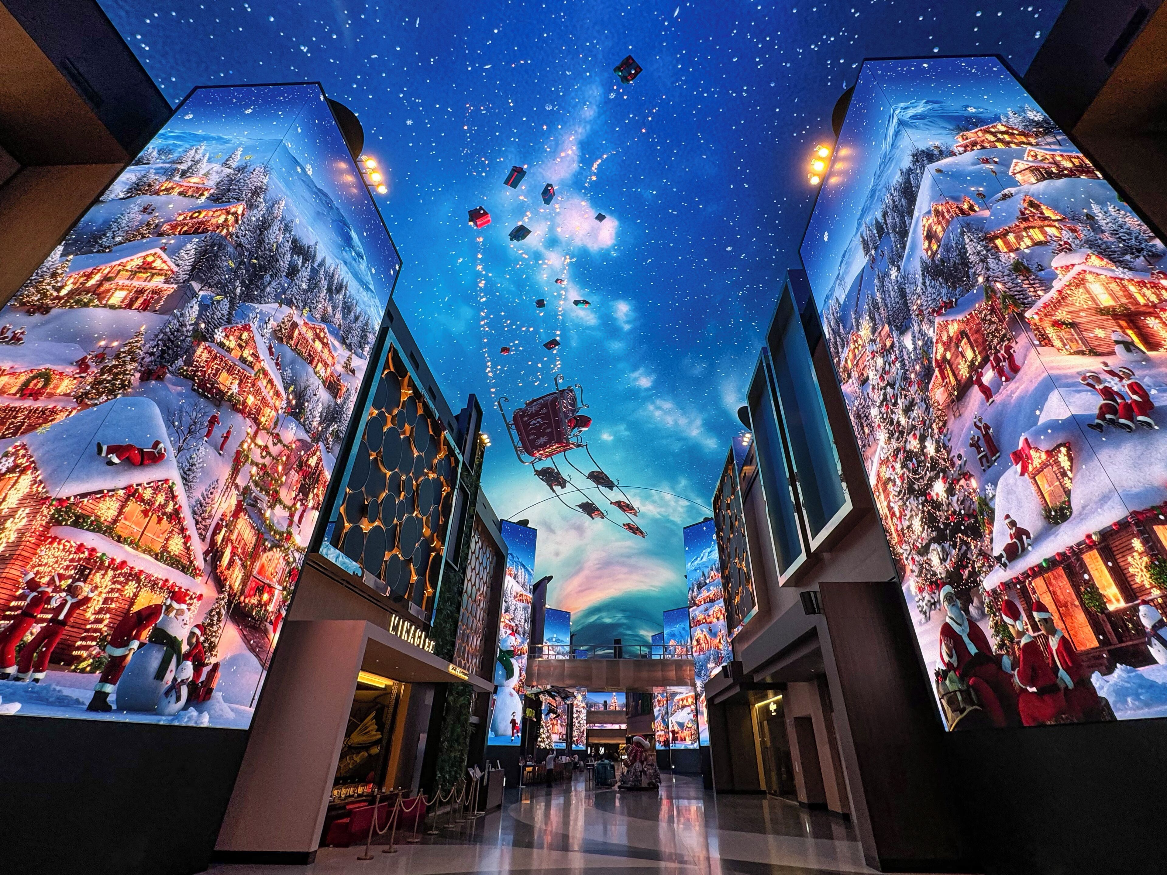 Un centro comercial con grandes pantallas digitales en las paredes que muestran una escena invernal festiva con nieve y adornos navideños.