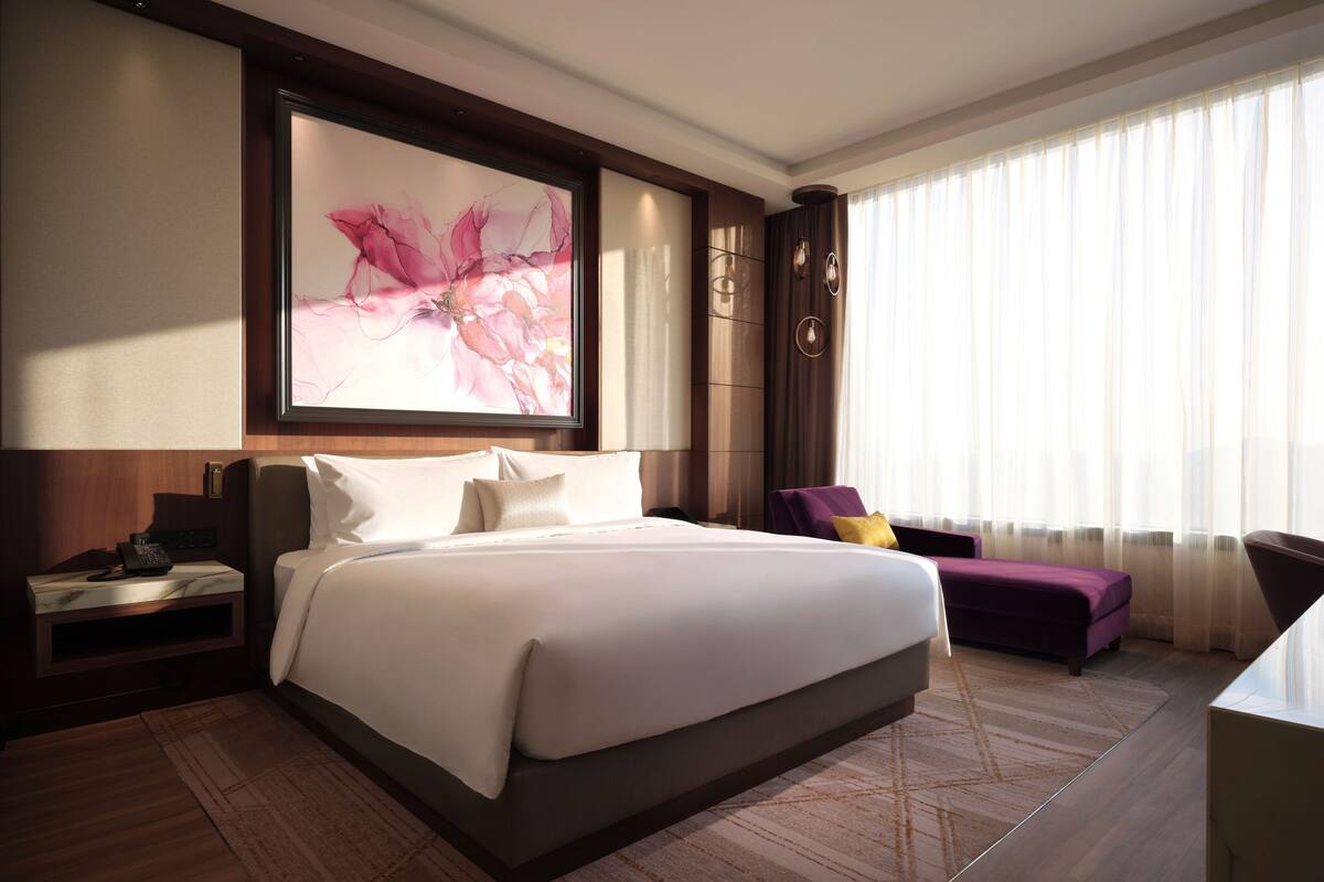 A modern hotel room with a large bed, a desk with a phone, a purple chair, and a large window with sheer curtains.