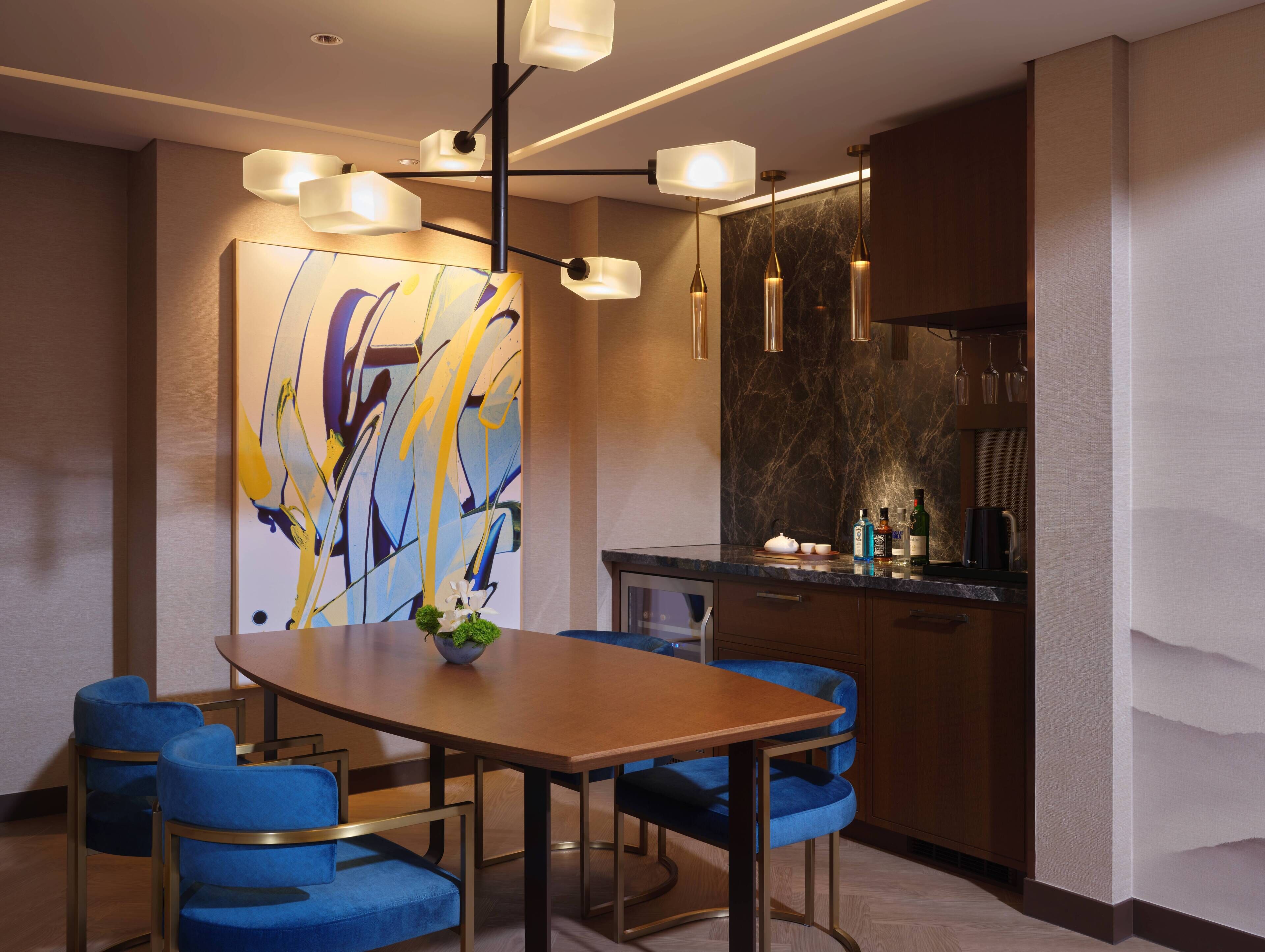 A modern dining area with a wooden table, blue upholstered chairs, a central vase, and a contemporary abstract painting on the wall.